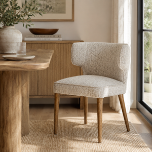 A modern upholstered dining chair with a textured light beige fabric and tapered wooden legs, positioned in a bright, contemporary dining room next to a wooden table.