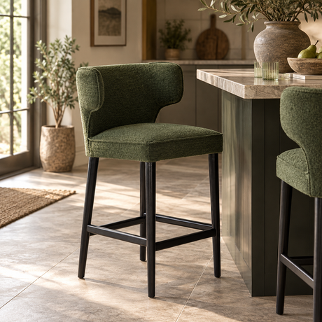 An upholstered bar stool in textured olive green fabric with black legs, positioned in a modern kitchen setting next to a stone-topped island.
