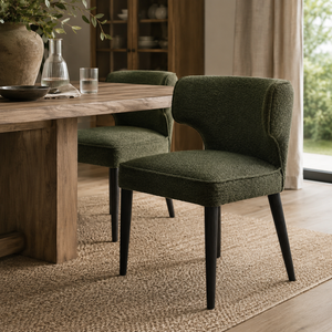 An upholstered dining chair in textured moss green fabric with black legs, shown in a real dining room beside a wooden table and rug.
