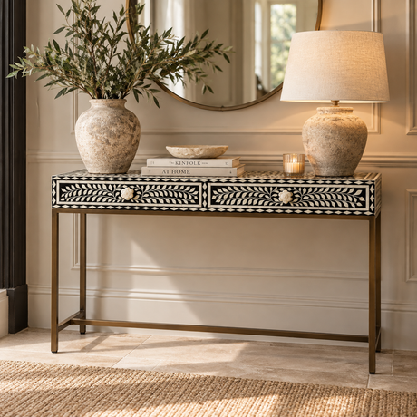 A bone-in-set console table with a brass frame, styled in a warm entryway with a vase of greenery, a lamp, books, and a candle on a light stone floor.