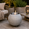 A spherical concrete outdoor fire pit with a visible flame, placed on a patio beside a wicker chair, plants, and a small table with a wine glass and olives.