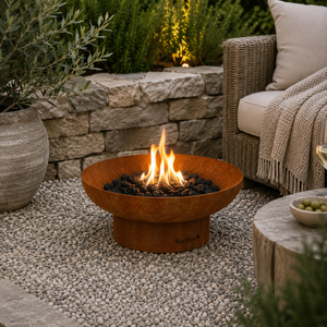 A round rusted metal fire pit bowl with a base, filled with black fire rocks and lit flames, placed on a pebble patio in an outdoor garden setting next to a wicker sofa.
