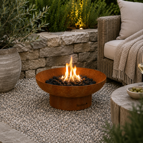 A round rusted metal fire pit bowl with a base, filled with black fire rocks and lit flames, placed on a pebble patio in an outdoor garden setting next to a wicker sofa.
