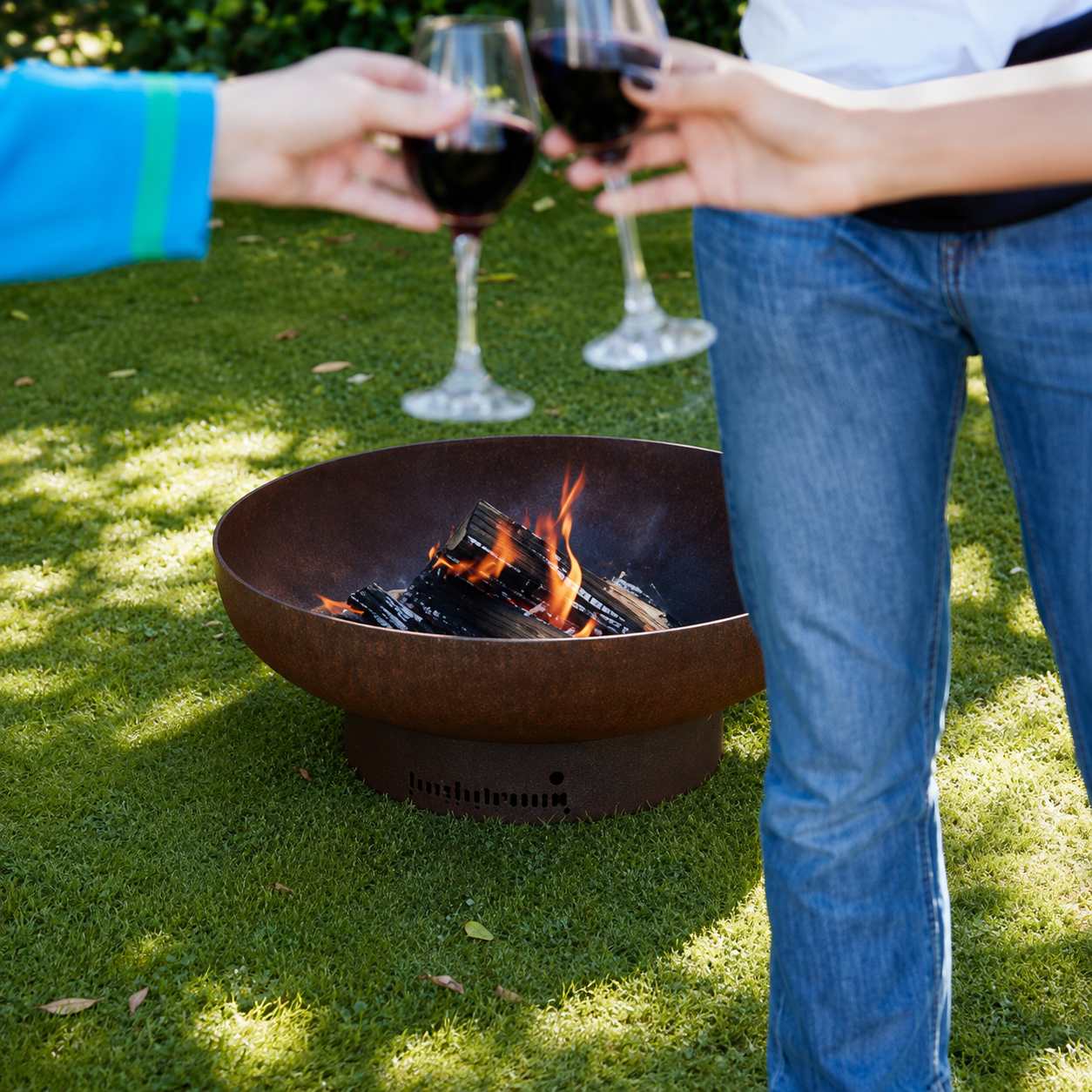 A round metal fire pit with visible flames sits on green grass outdoors, while two people hold wine glasses in the background.