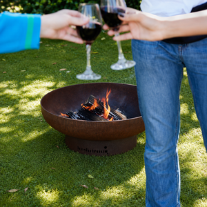A round metal fire pit with visible flames sits on green grass outdoors, while two people hold wine glasses in the background.