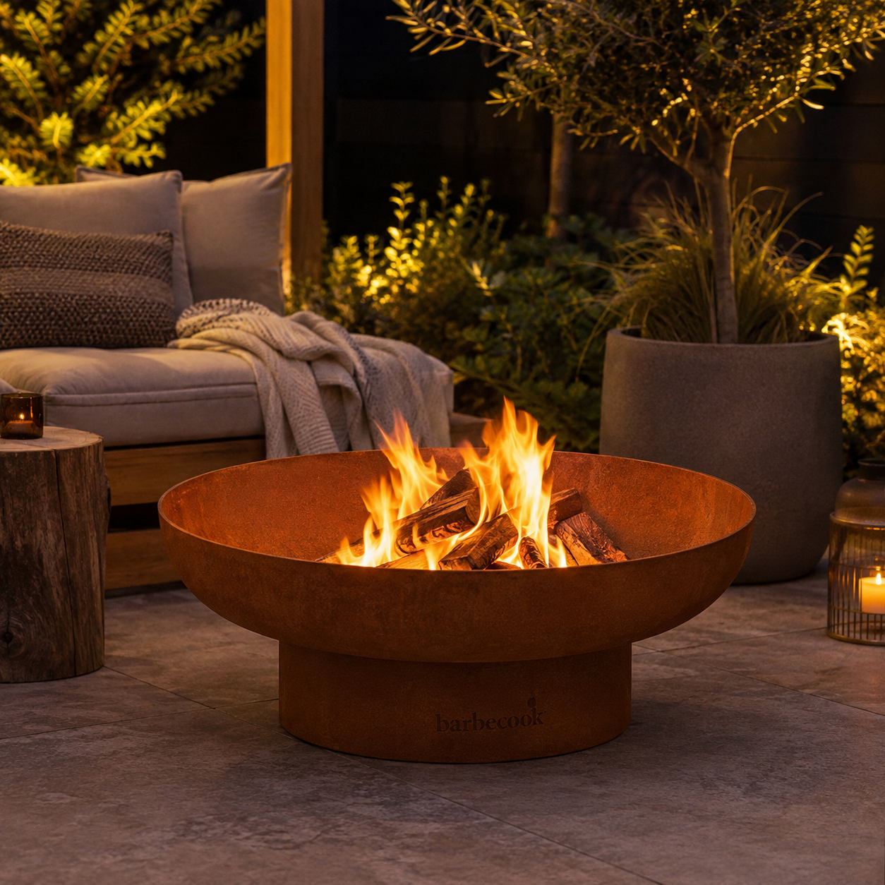 A round outdoor fire pit bowl with a base, shown lit with orange flames on a patio. The product has a rusted orange-brown metal finish and is branded 'barbecue' on the front.