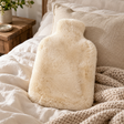 Beige Sheepskin Hot Water Bottle resting on a linen bed with a chunky knitted throw, beside a rustic wooden bedside table
