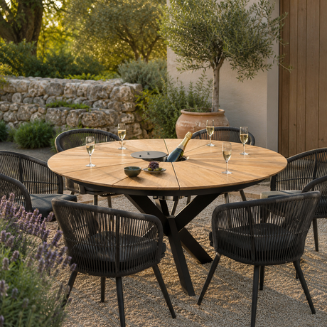 Outdoor round dining table set with black woven chairs in a patio garden setting, with wine glasses, a bottle, and a center tray on the tabletop.