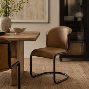Tan leather dining chair with a curved back and black metal cantilever frame, positioned beside a rustic wooden dining table in a warm, neutral-toned interior with a woven rug and natural decor.