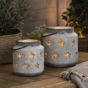 Two rustic grey lanterns with star-shaped cutouts, lit with warm yellow-orange fairy lights inside, displayed on a wooden table with potted plants in a cozy outdoor setting.