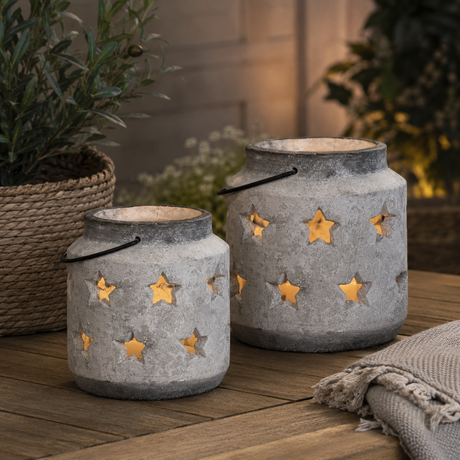 Two rustic grey lanterns with star-shaped cutouts, lit with warm yellow-orange fairy lights inside, displayed on a wooden table with potted plants in a cozy outdoor setting.