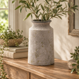 Bloomville Urn Stone Vase filled with olive branches on a wooden sideboard by a window, beside books and a trailing plant