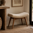A low upholstered bench with a curved cream bouclé seat and medium brown wooden legs, placed in a warm neutral room beside a wooden table and framed wall art.