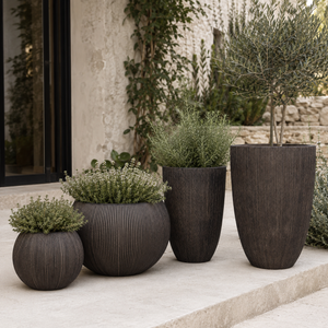 Four dark brown ribbed planter pots of varying sizes arranged on a light stone patio in front of a textured stucco wall with climbing greenery.