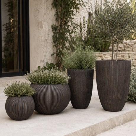 Four dark brown ribbed planter pots of varying sizes arranged on a light stone patio in front of a textured stucco wall with climbing greenery.