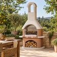 Buschbeck Toscana Masonry Barbecue in white quartz and terracotta with food on the grill and a log store full of firewood, on a sunlit stone terrace beside a rustic dining table and olive trees
