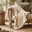 Cappuccino Merino Wool Blanket in cream with a caramel binding draped over a wooden armchair, beside a stone vase of olive branches and a jute rug