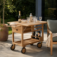 A light natural wood rolling bar cart with wheels, shown on an outdoor patio. The cart is styled with a bottle, glasses, a shaker, and food on its top surface and open shelves.