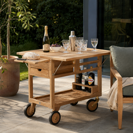A light natural wood rolling bar cart with wheels, shown on an outdoor patio. The cart is styled with a bottle, glasses, a shaker, and food on its top surface and open shelves.