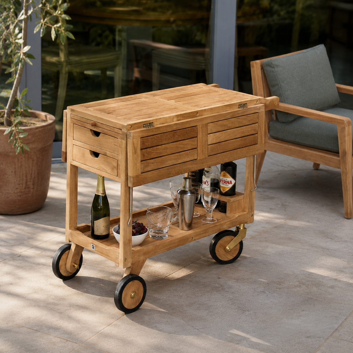 A light natural wood rolling bar cart with drawers and a lower shelf, styled on a patio with a chair and potted plant in the background.
