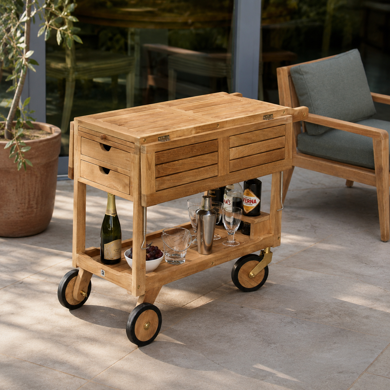 A light natural wood rolling bar cart with drawers and a lower shelf, styled on a patio with a chair and potted plant in the background.