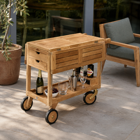 A light natural wood rolling bar cart with drawers and a lower shelf, styled on a patio with a chair and potted plant in the background.