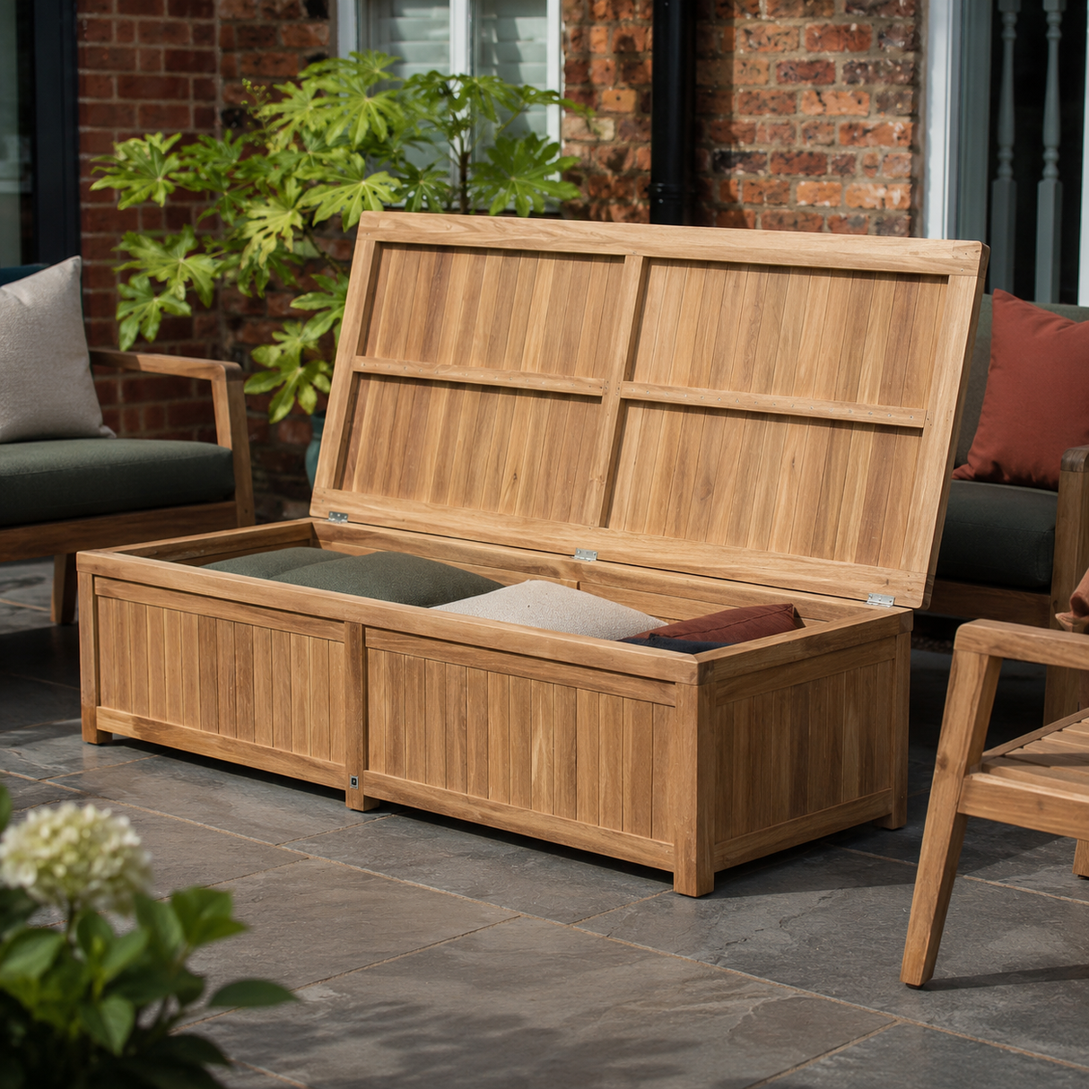 A wooden outdoor storage bench with the lid propped open, showing cushions inside, placed on a stone patio in front of a brick house with patio furniture nearby.