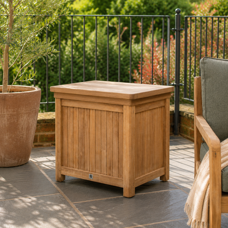 A rectangular wooden outdoor storage chest with a slatted front and flat lid, shown on a patio next to a chair with a blanket, with a garden and railing in the background.