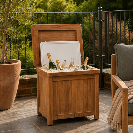 A wooden outdoor storage cooler with its lid open, placed on a patio with bottles and ice inside. The cooler is made of warm medium-brown wood with a white lid insert, set against a background of green foliage and a black metal fence.