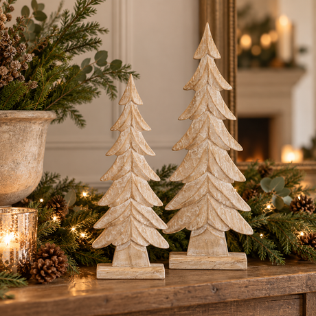 Small and large whitewashed carved wood trees on a mantel with pine greenery, fairy lights and a candle