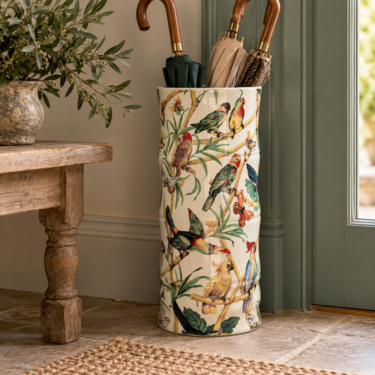 A tall umbrella stand with a cream background and a colourful bird-and-branch print, holding several umbrellas, placed indoors near a wooden table and door.