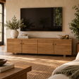 A mid-century modern wooden TV stand and sideboard cabinet in a warm natural wood finish, styled in a living room setting with a wall-mounted television, potted plants, and woven decor.