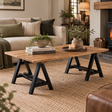 A rectangular wooden coffee table with black trestle-style legs in a cosy living room setting. The table is styled with books, a small potted plant, and a candle on a round tray, set on a woven jute rug in front of a sofa.
