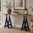 A rustic console table with a medium brown wooden top and black trestle-style legs, styled in a home interior with a potted plant, books, a lantern, framed wall art, and a woven basket.