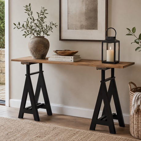 A rustic console table with a medium brown wooden top and black trestle-style legs, styled in a home interior with a potted plant, books, a lantern, framed wall art, and a woven basket.