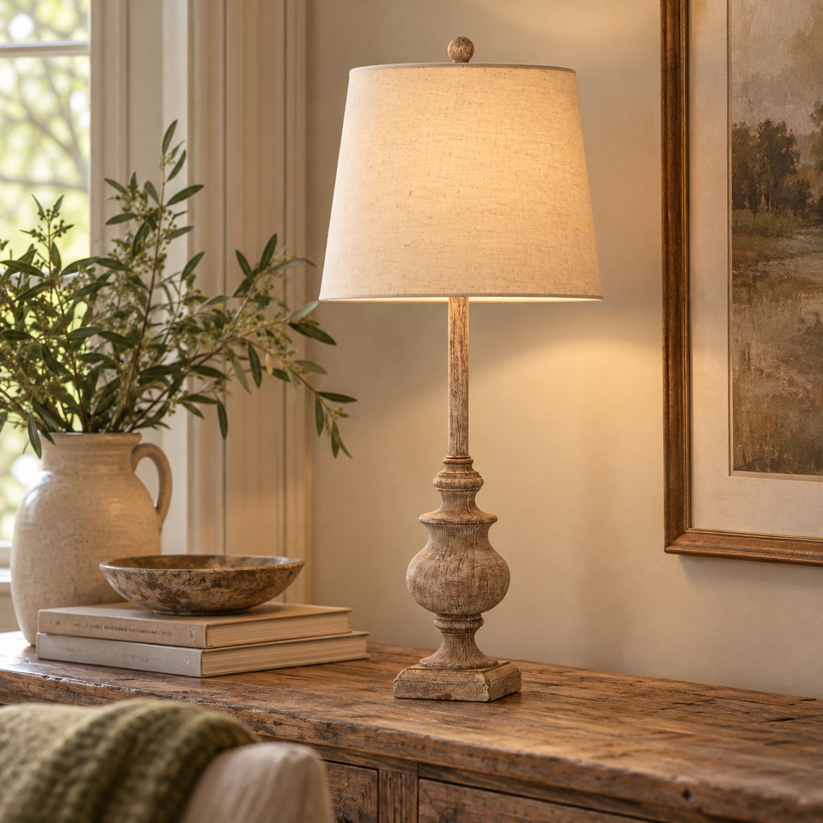 Chiaro Candlestick Table Lamp With Linen Shade lit on a rustic wooden console with olive branches in a stoneware jug