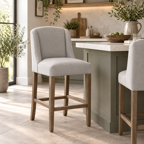Light grey upholstered bar stool with a curved back and natural wood legs, styled beside a kitchen island in a bright, neutral interior.