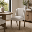 An upholstered dining chair in a light beige fabric with tapered wood legs, shown in a styled dining room next to a round wooden table and a sideboard.