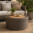 Round outdoor wicker ottoman table with a light wood top, styled in a patio setting with a sofa, pillows, a potted fern, a bowl of olives, and a glass of iced tea.