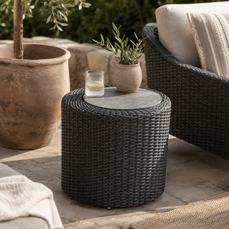 A dark gray woven round side table with a circular stone top, styled on an outdoor patio with a glass candle holder and a small potted plant on top.