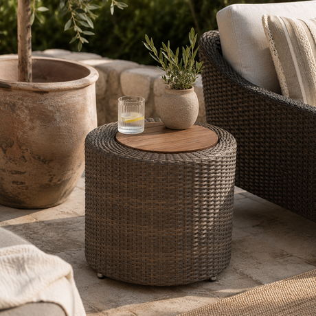 A round woven wicker side table with a light wood top, styled on a stone patio next to outdoor furniture. The table holds a small glass candle holder and a small potted green plant.