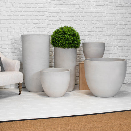 Five light gray concrete planters in various shapes and sizes arranged on a white wooden floor against a white brick wall, with a green topiary in one planter and a light beige armchair partially visible on the left.