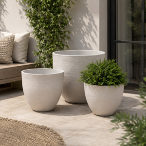 Three textured off-white planter pots in different sizes arranged on a patio, with one pot holding a green shrub. The setting includes a bench with pillows, a climbing plant, and a glass door in a real outdoor environment.