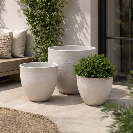 Three textured off-white planter pots in different sizes arranged on a patio, with one pot holding a green shrub. The setting includes a bench with pillows, a climbing plant, and a glass door in a real outdoor environment.