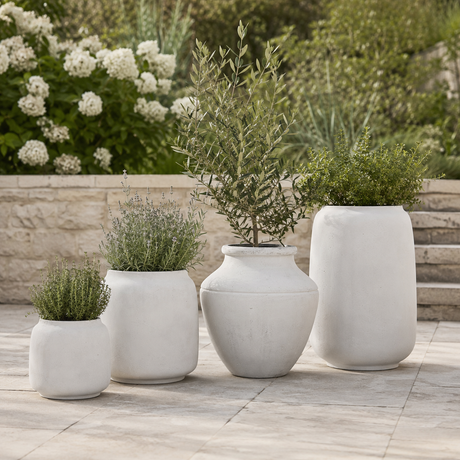 Four white off-white planters in graduated sizes arranged on a stone patio with green plants and a garden background.
