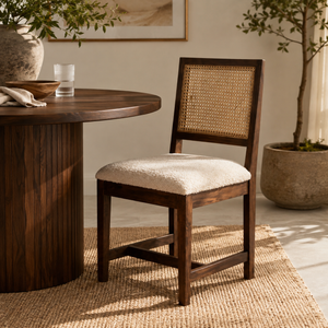 A wooden dining chair with a woven cane backrest and a cream bouclé seat cushion, styled next to a round wooden table on a jute rug in a warm, sunlit room.