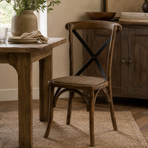 A rustic wooden dining chair with a woven seat and black cross-back support, shown in a styled dining room next to a wooden table.