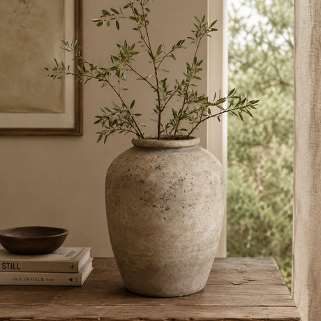 Large terracotta vase with green branches on a wooden surface, with a window and painting in the background.