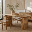 A rustic rectangular wooden dining table with thick block-style legs, surrounded by matching wooden chairs with woven seats, in a bright, modern dining room with a vase of greenery and a bowl on the table.