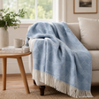 A blue wool blanket with a diamond pattern and a cream fringe draped over a beige sofa beside a side table with books and a coffee cup